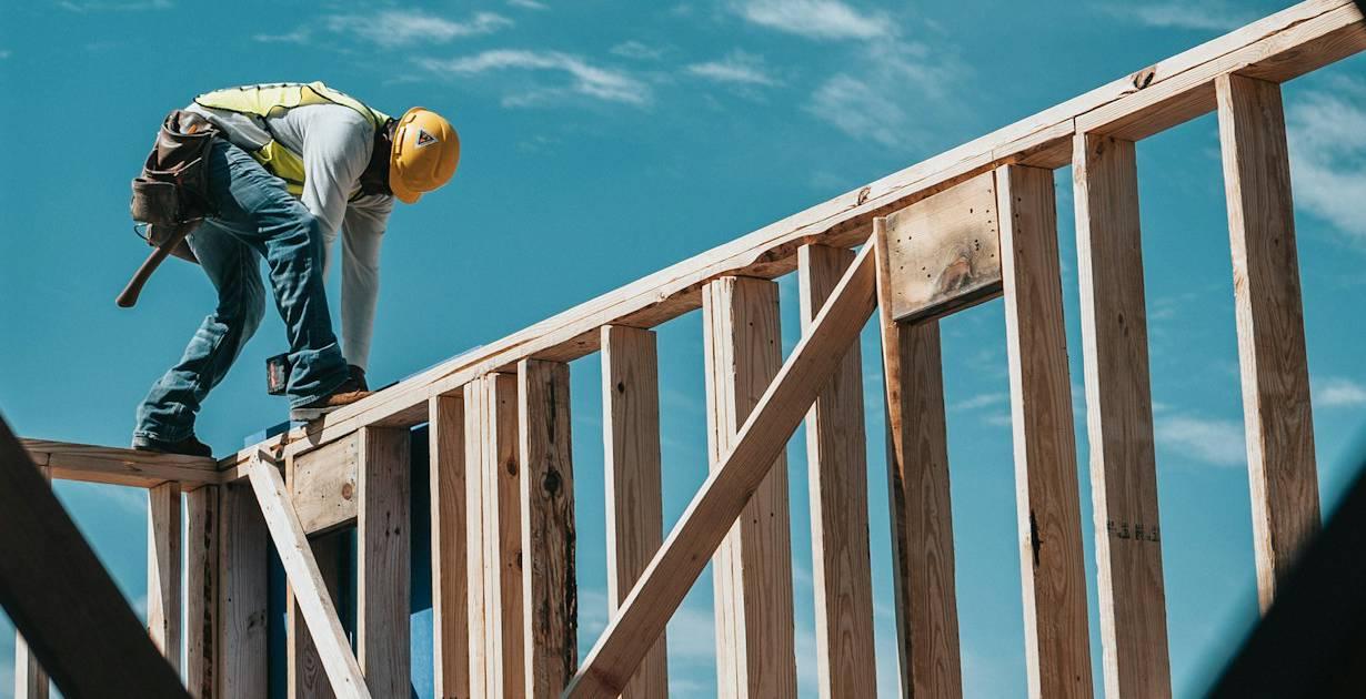 Construction worker wearing a yellow hard hat building a wooden frame under a clear blue sky.